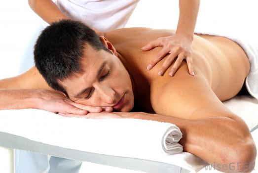 man with black hair lying on a white massage table with a woman's hands massaging his shoulders